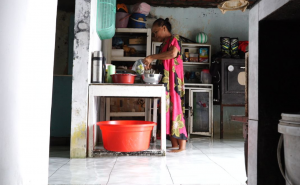 Alfiyah, 52 tahun, Ketua RT 05 RW 16 Tambak Rejo, tengah mengumpulkan bahan makanan di dapurnya yang terendam air rob. Dia berpindah ke teras rumahnya—satu-satunya bagian rumahnya yang kering untuk menyiapkan makan siang, Alfiyah mengaku ingin pindah, tapi karena mengutamakan pendidikan putranya yang tengah kuliah, niat itu mesti ia tunda. (Foto: Eka Winarotomo)